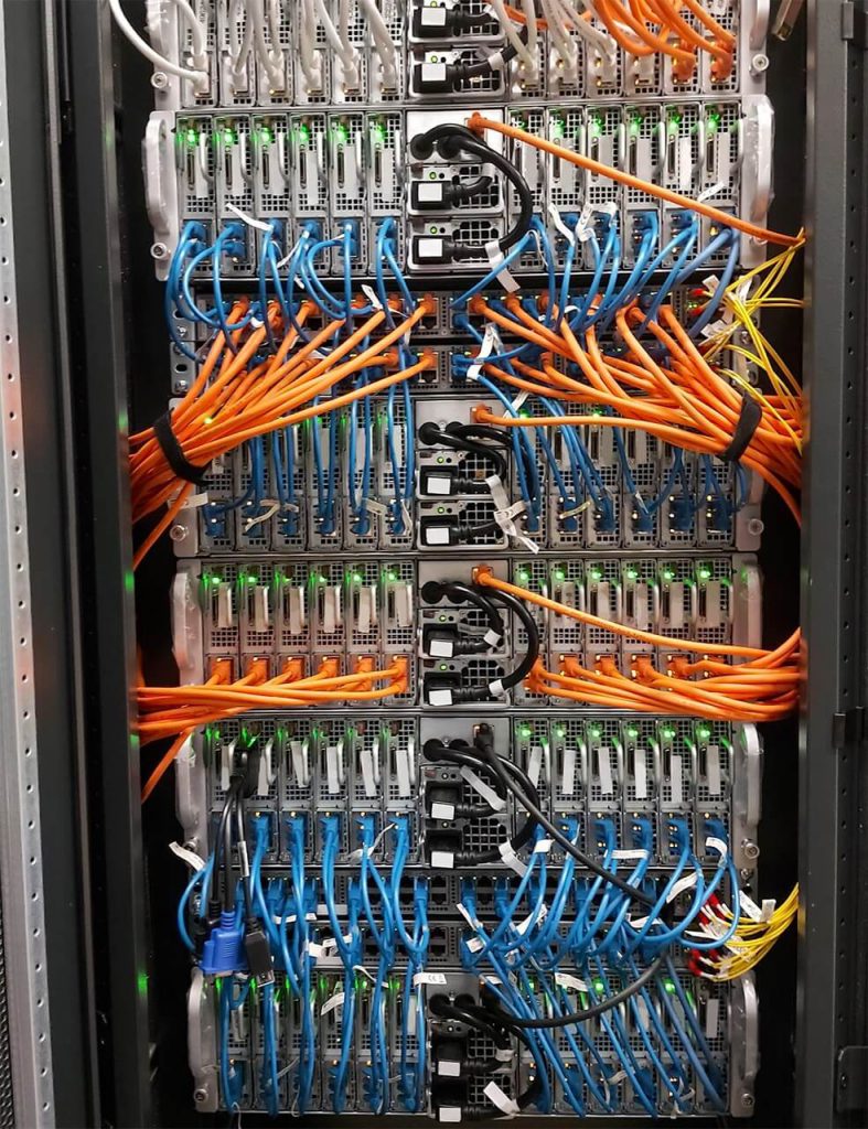 Server rack with multiple network switches connected by orange, blue, and yellow cables. Green indicator lights are visible.