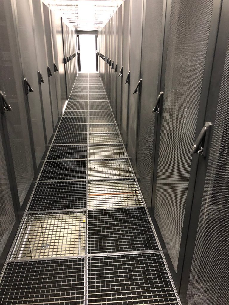 Narrow corridor with metal grating floor, flanked by tall server racks, leading to a bright doorway at the end.